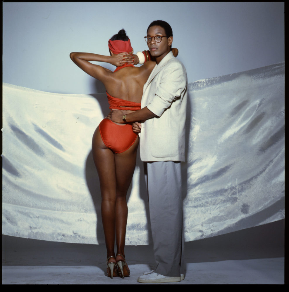 Photograph of dark skin man in white blazer and gray trousers holding waist of a dark skin woman posing next to him in an orange bathing suit
