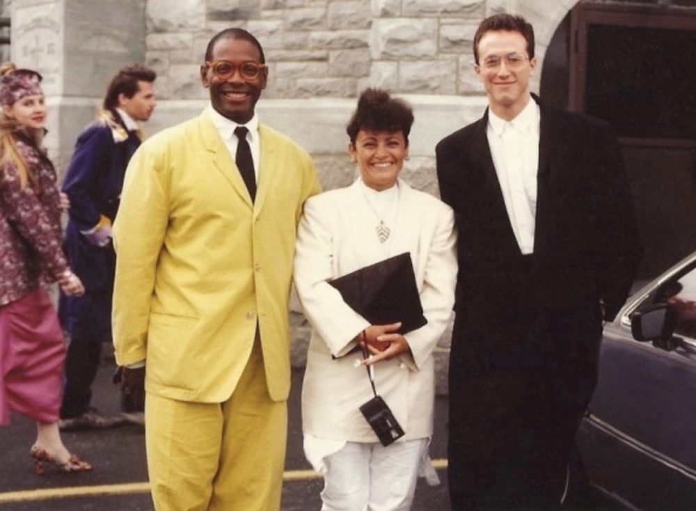Two men and a woman stand smiling in front of a stone building facade. The young Black man on the left wears a bright yellow suit, the woman in the middle wears a white suit and holds a black bag and a camera hanging from her wrist, and the young white man on the right wears a black suit and white shirt.