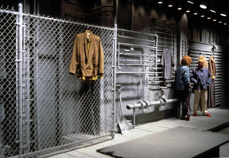 Image of two models talking in the WilliWear New York Showroom, surrounded by a gray chain-link fence, gray window-bars, and wooden planks which WilliWear garments hang from