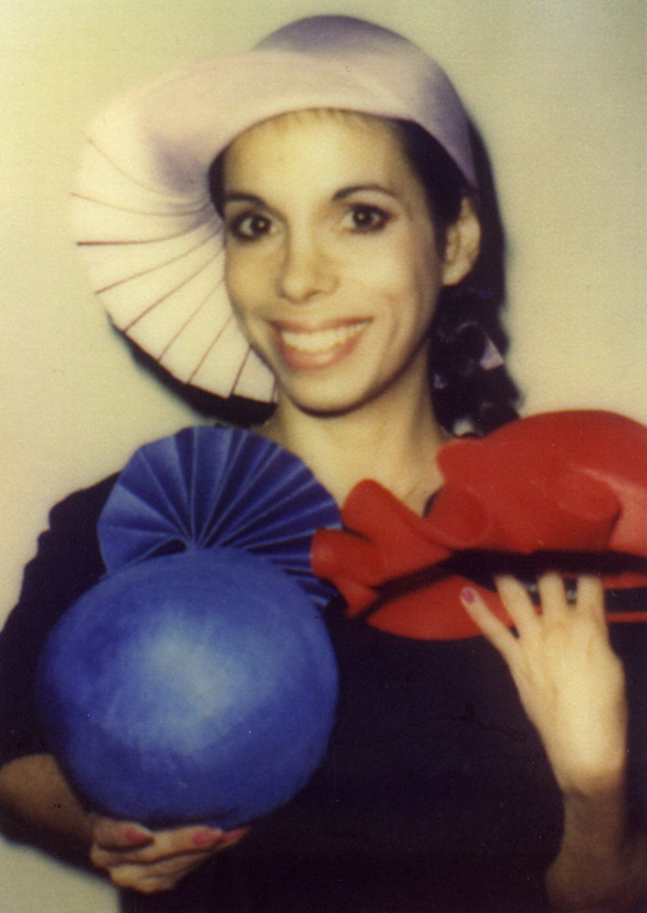 Color photograph of a woman smiling at the camera. She wears a white pleated had. In her right hand she holds a blue hat with flowering pleats, and in her left an ornate red hat.