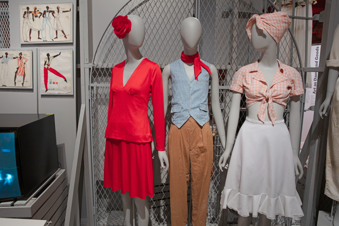 In this scene from the exhibition, three mannequins are propped in front of chain link fence painted a light grey, in front a wall painted all grey. On the left, the mannequin wears matching red top and skirt with red flower perched on the right side of her head. In the center, the mannequin wears a blue and white striped vest and brown pants with a red scarf. On the right, the mannequin wears a white skirt and pink checkered top and scarf tied on her head.