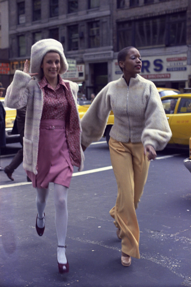 Color photograph of two woman running to cross a busy New York Street. They are smiling, holding hands as they run.
