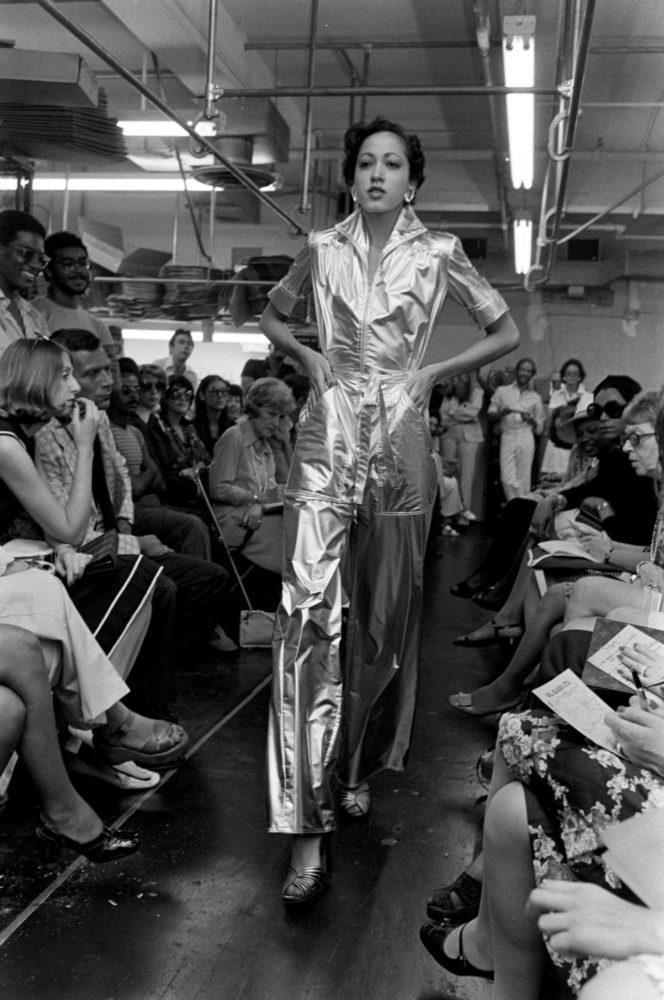 Grayscale photograph of a model walking confidently down a runway in a chrome jumpsuit with hands in pockets. A room full of men and women, seating and standing, look at the model as she walks.