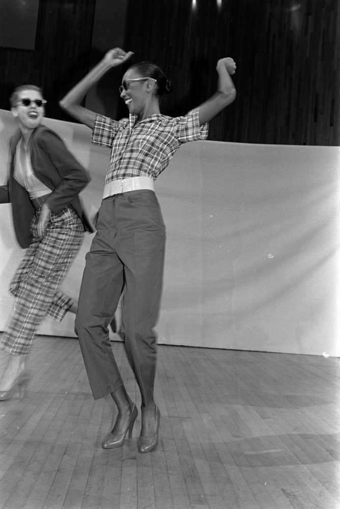 A greyscale photo. A light-skinned dancer and a dark-skinned dancer smile and twist in front of a white curtain on stage. They are dressed oppositely, with the figure on the right wearing a plaid top and grey pants, and the other wearing a grey suit top and plaid pants.
