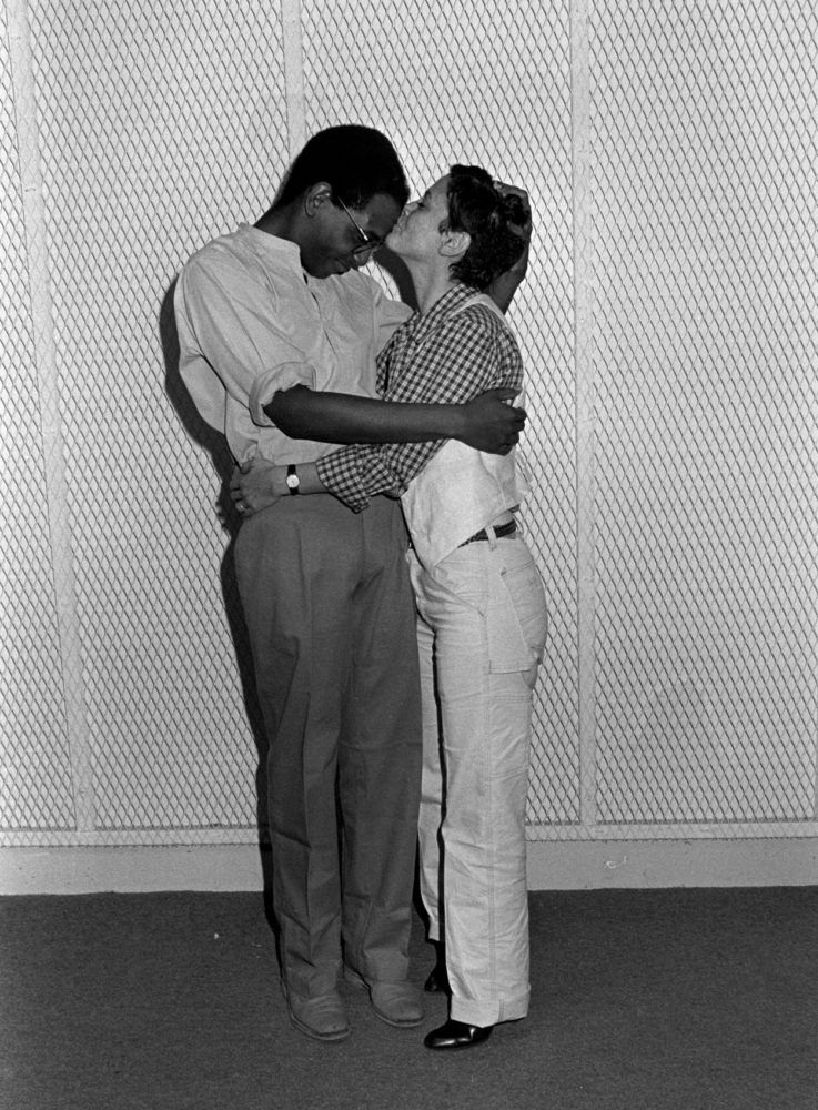 Grayscale photograph of a Black man with round glasses and a white woman embracing in a hug; the woman kisses the man on the head