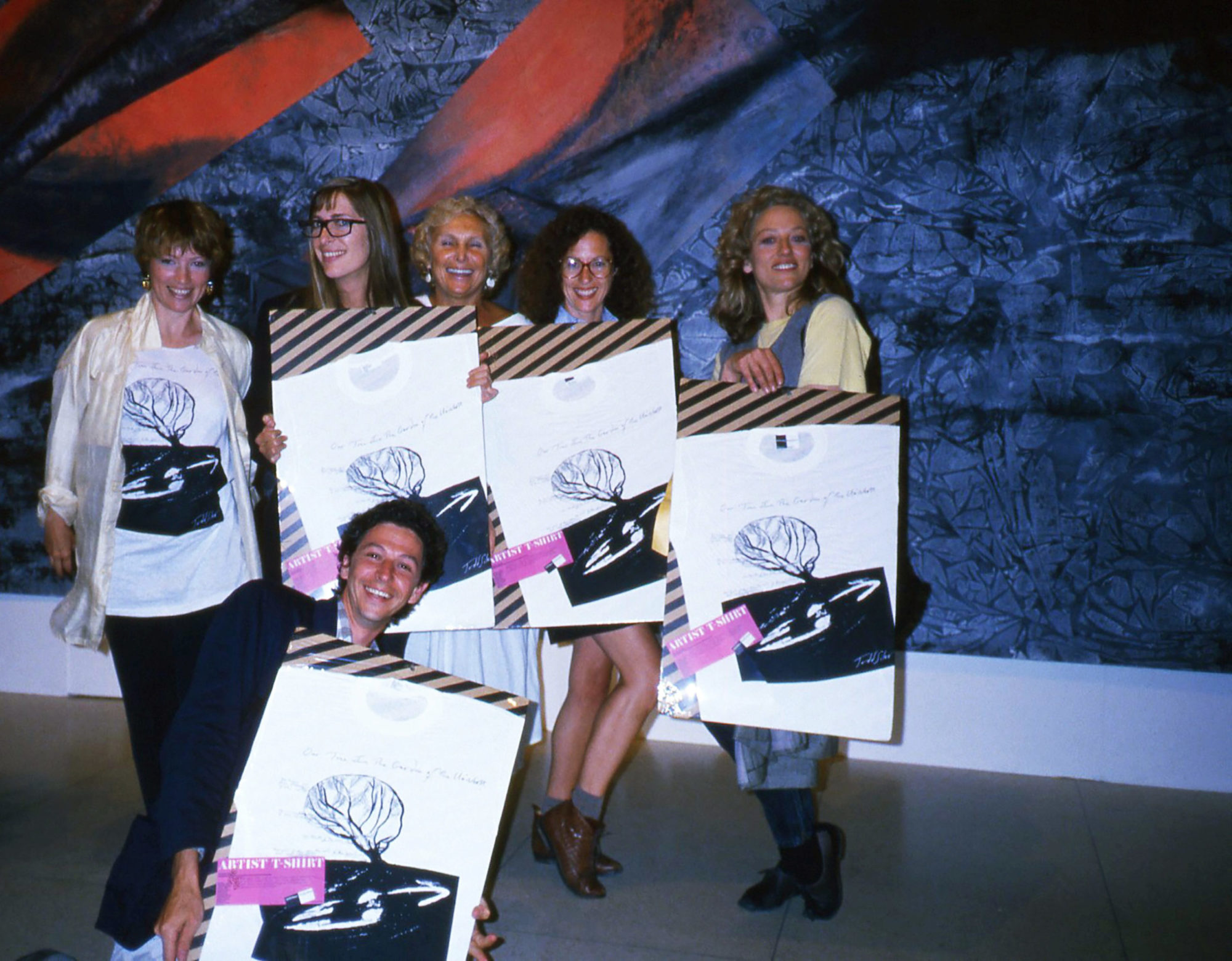 A color photograph of a group of people holding signs. Five women stand holding posters with the design of the white tshirt, while one standing on the left wears the shirt. A man kneels in front holding another sign. The poster shows the white shirt on a striped background and over a purple box it reads "Artist T-Shirt".