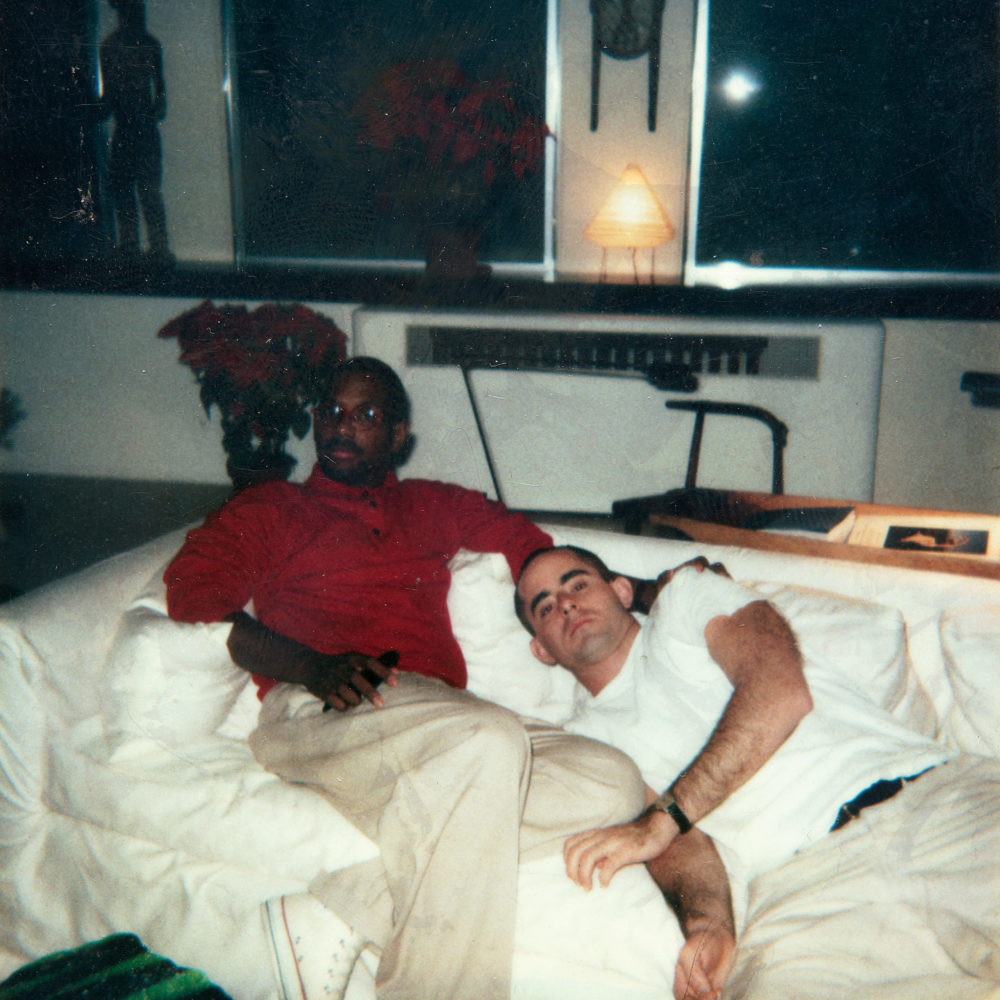 Photo of two men lounging on a white sofa in a white room with black curtains, African art, and poinsettias