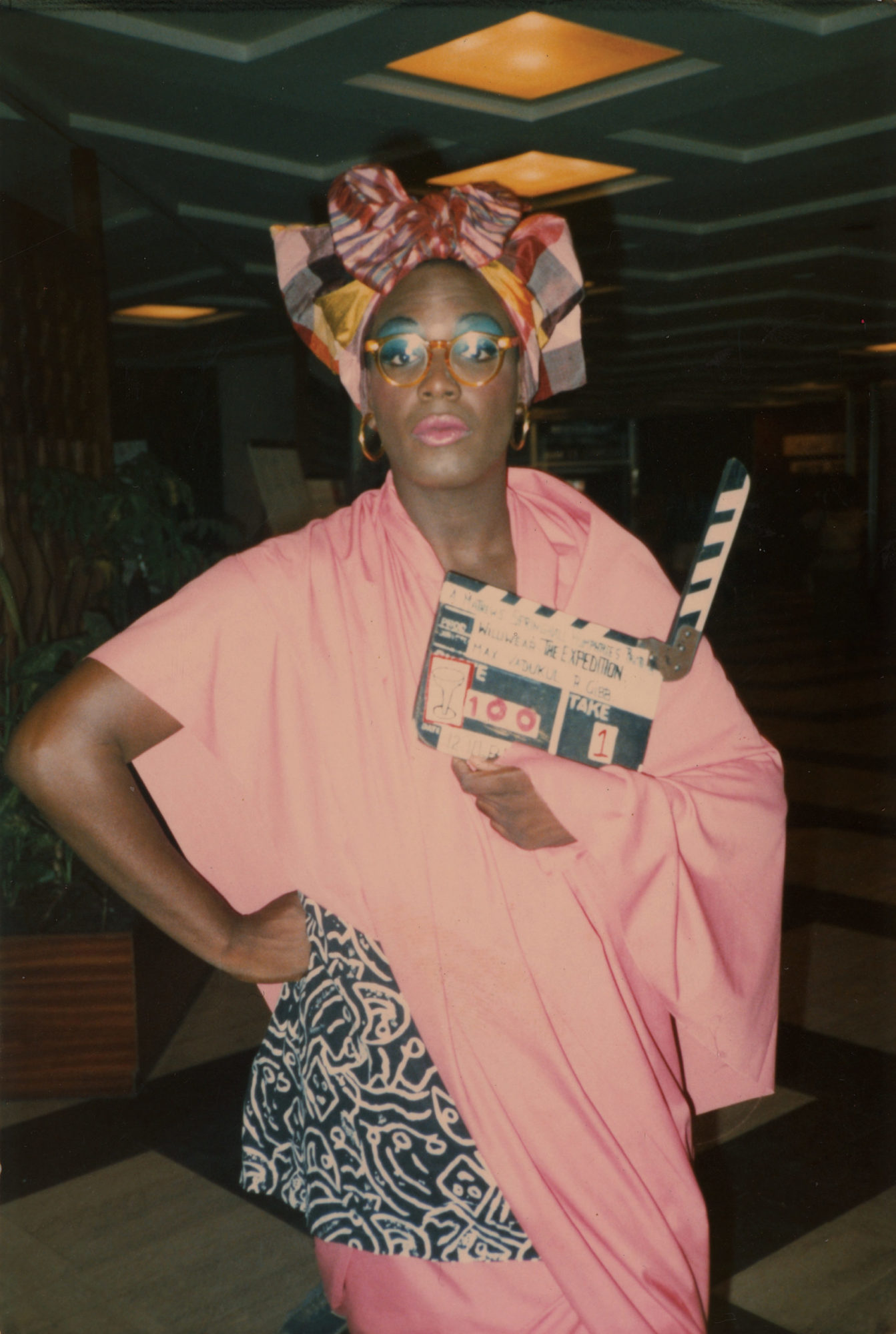 Photograph of a bespectacled dark-skinned Black man, Willi Smith, dressed in a pink and black women's ensemble, wearing colorful makeup, a headwrap, and holding a film slate