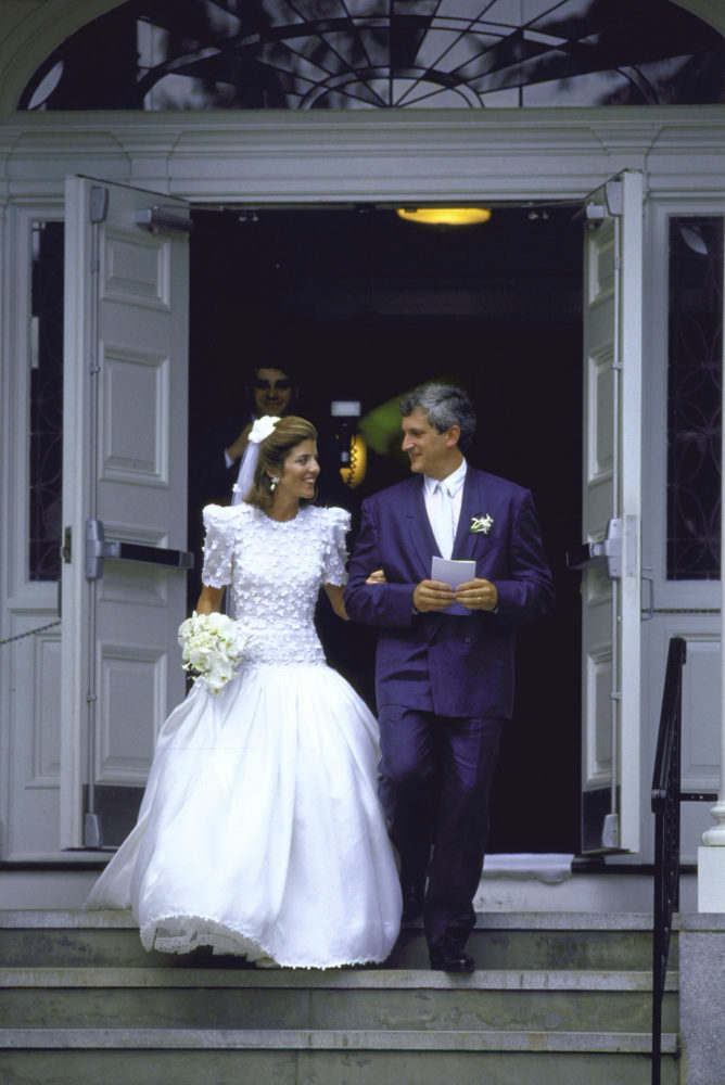A man and a woman walk out of a church, looking at each other. She wears a long white wedding dress with white flowers on top, holding a bouquet, and he wears a blue suit. A photographer stands in the darkened doorway behind them.
