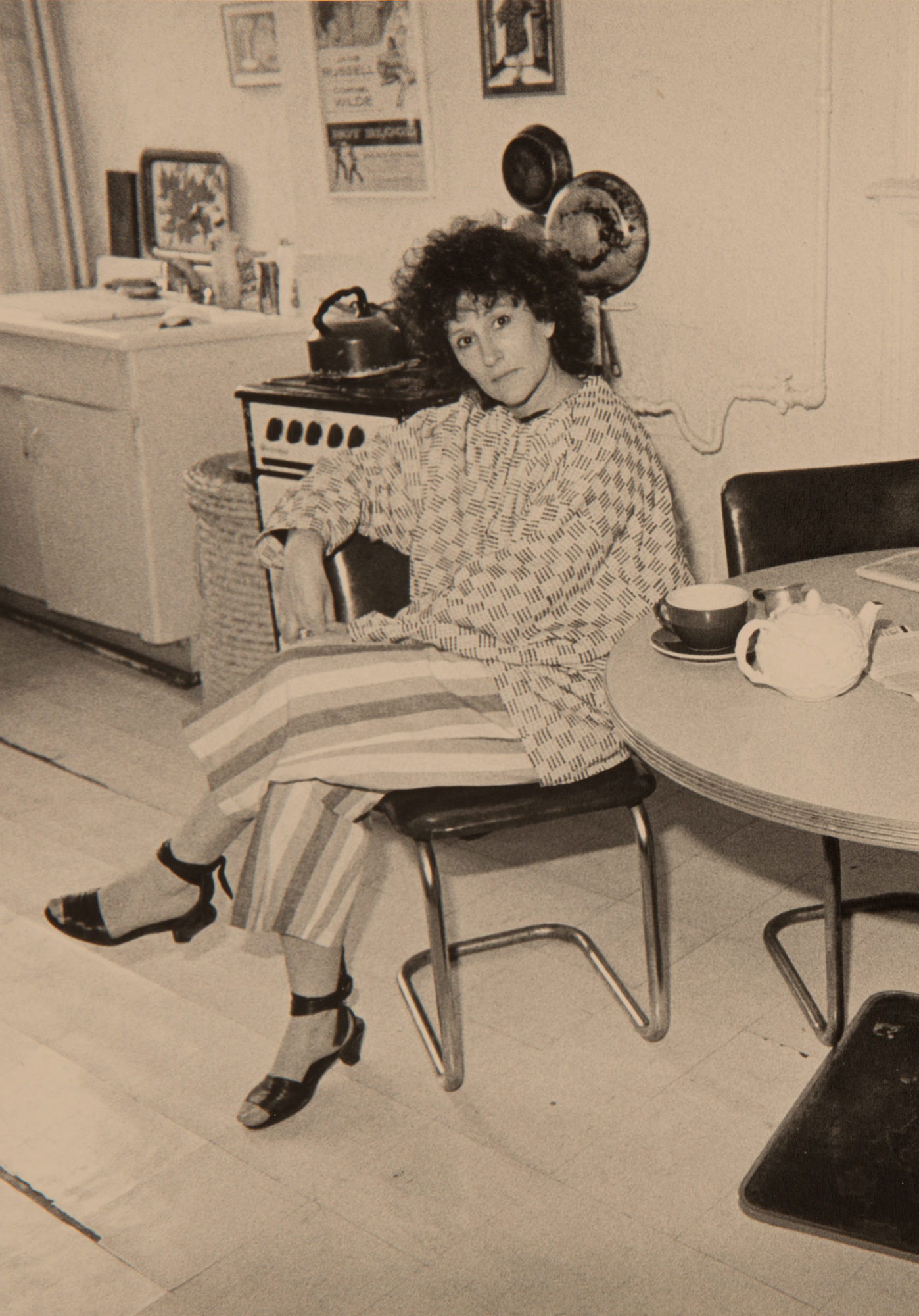 Sepia photo of a woman dressed in a patterned WilliWear jacket and striped pants sitting in a kitchen