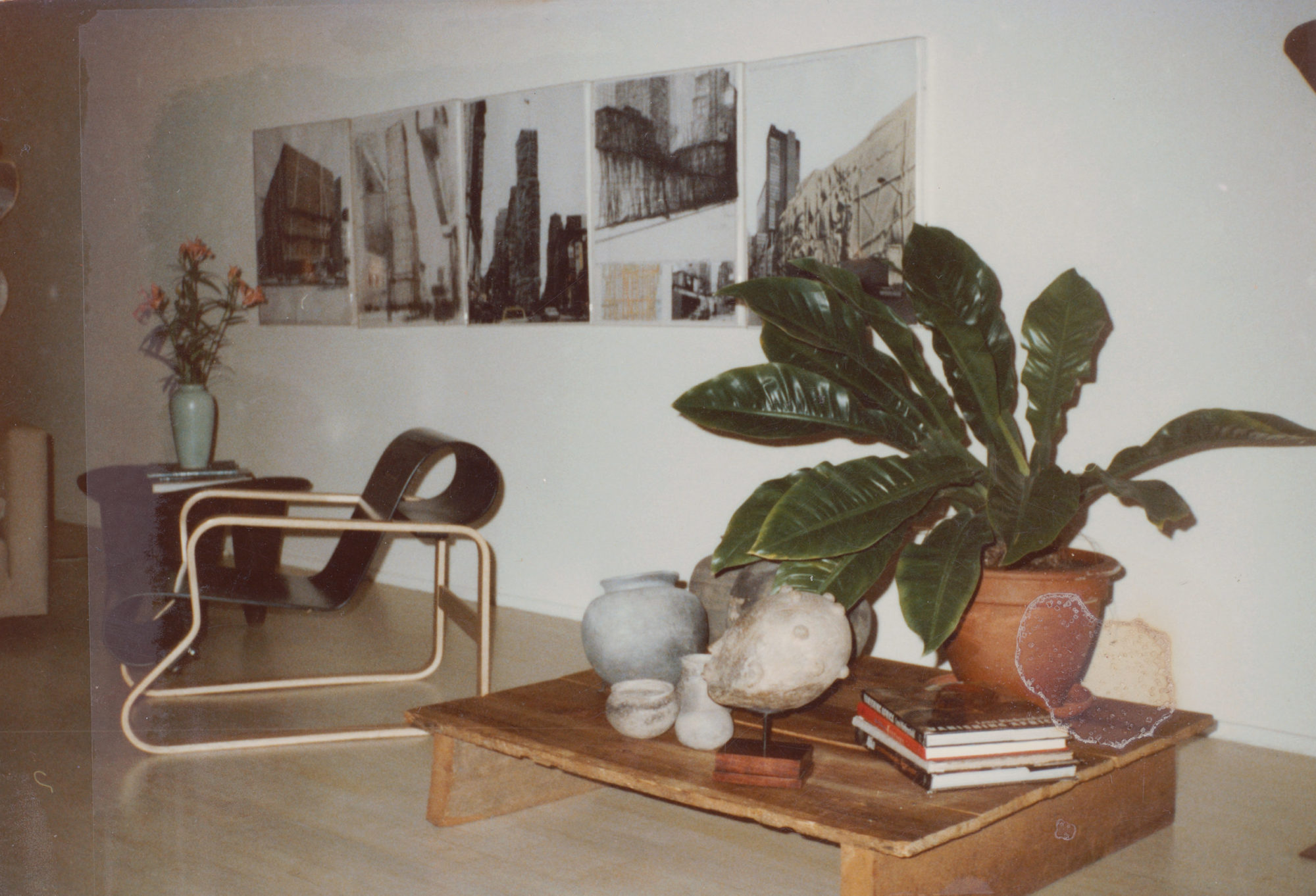 An interior photograph of a home. A low table sits on the floor on the right of the image. A large plant, books, pots, and sculpture of a figure's head sit on the table. To the left of the table is a geometric chair. On the wall behind it is a row of large drawings of buildings.