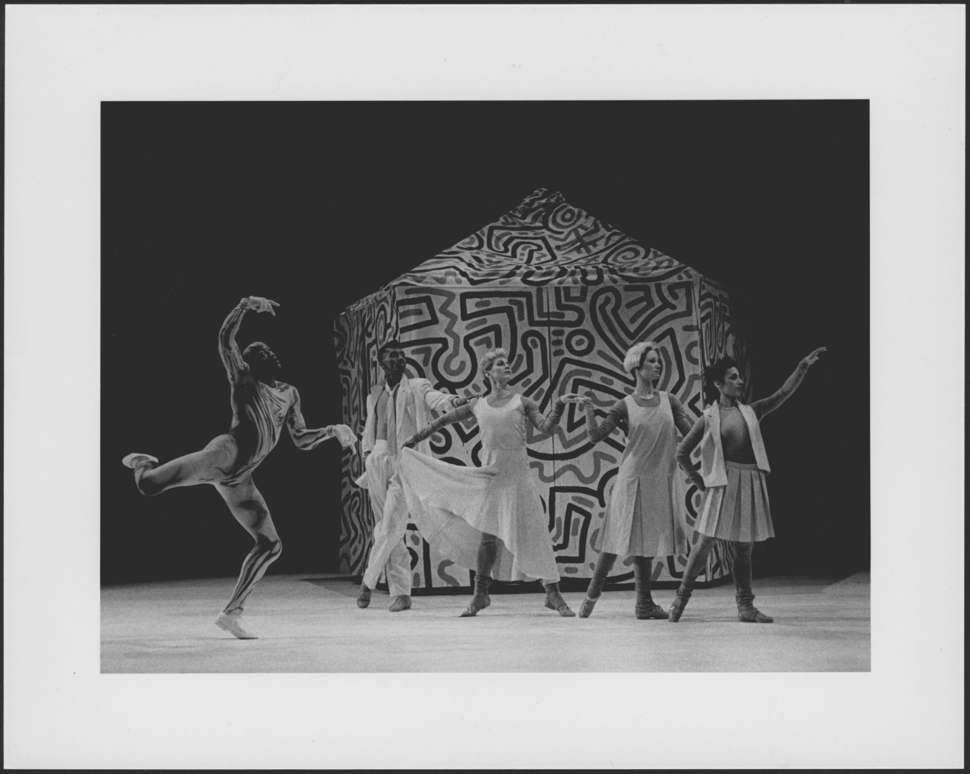 Grayscale photograph of five dancers on stage posing in dramatic manner with patterned tent in the background