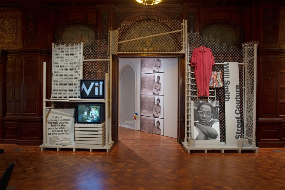 Installation view for the Willi Smith: Street Couture exhibition in the Great Hall at Cooper Hewitt showing items on grey chain-link fencing, wooden pallets, and industrial piping. On the left of the entrance is a gray folding security gate and box television with WilliWear graphics projected resting on wooden planks. Below these items are another box television displaying a fashion presentation and a board with text reading, [I don’t / design clothes / for the queen / but for the / people who / wave at her / as she goes by.] On the right of the entrance, a grayscale portrait of a dark-skinned man with round glasses (Willi Smith) rests against a gray metal ladder and chain link fence. To the right of the portrait, a long board of printed text reads, [/Willi Smith/ Street Couture/]. Beside the board, a pair of multicolored shorts and a long red nylon coat hang from the chain-link fence.