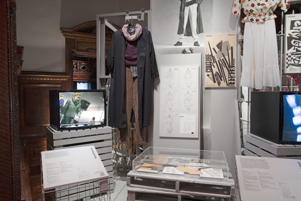 Installation view of the Willi Smith: Street Couture exhibition. The image shows a box television, six illustrations from the WilliWear Street Couture collection, a large grayscale publicity portrait of a model wearing garments from the collection, and an ensemble composed of a lavender scarf, plum sweater, button-up striped shirt, long brown pants, and a long black coat. To the right of this display to a second display showing part of a long white skirt and a white blouse with colorful squares hanging beside a box TV and a black-and-white poster the reads, [City Island].