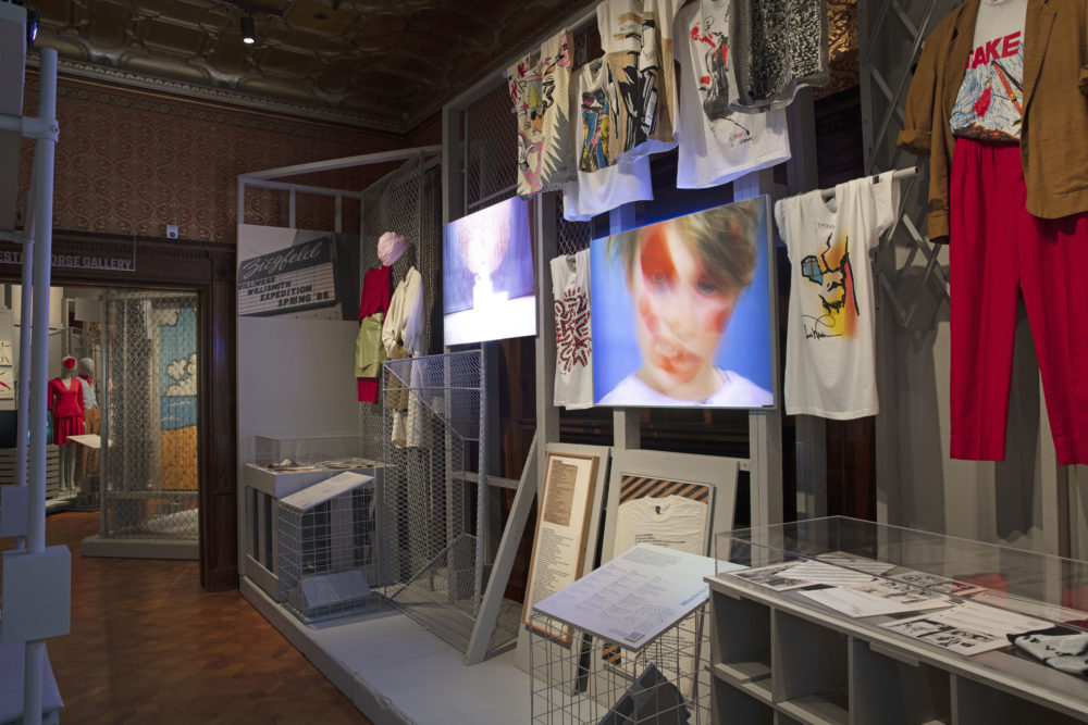 Seven graphic T-shirts are displayed hanging in a T form on metal rods. Between two graphic T-shirts, a projection board displays a film showing a young person with dramatic face paint. Sheets of paper displaying images and text related to the T-shirts and film are encased along with a graphic T-shirt to the right of the projected film. Below the projection board are two framed objects; one white T-shirt with screen printed black text and two pieces of paper bearing the same text. To the far left of this installation is another installation that shows two ensembles hanging from a gray chain-link fence beside another projection board that displays a different film.