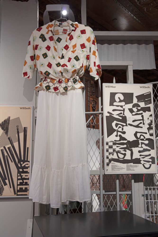 A long white skirt and a white blouse with multicolored squares hang in a wood-paneled room beside a box TV and a black-and-white poster the reads, [City Island].