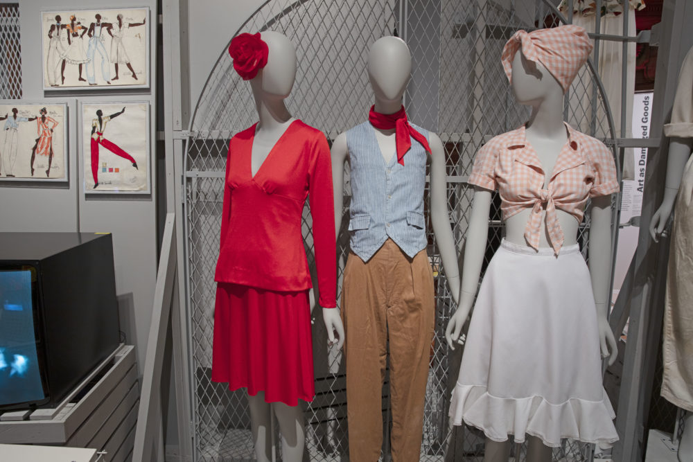 Three gray mannequins wearing clothing, face us. The mannequin on the far left wears a bright red long sleeve shirt with a V-neckline and a knee-length red skirt; a red flower is pinned onto the figure’s head. The mannequin in the center wears a light blue button-up vest with pockets, a red neck scarf, and long brown pants. The mannequin on the right wears a pink and white gingham headwrap, a tied pink and white gingham shirt with rolled short sleeves, and a white knee-length skirt. Beside the three manikins are fashion illustrations of the garments from the same production, Deep South Suite.