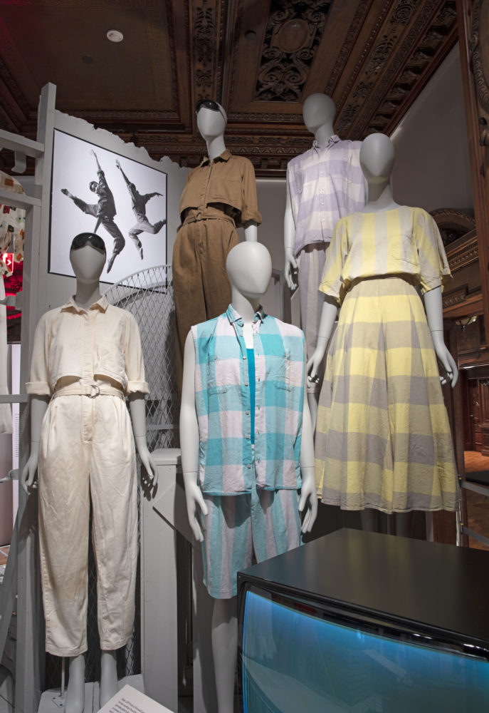Five gray mannequins are positioned together on a two-tiered platform. Three mannequins are on the lower platform, facing us; two are on a higher platform behind the three and are positioned at three-quarter view. Two mannequins wear aviator jumpsuits in neutral colors with black goggles. Three mannequins wear plaid garments in white and gray, yellow and gray, and blue and white. A grayscale photograph of two dancers wearing the same jumpsuits and goggles is on display beside the mannequins.
