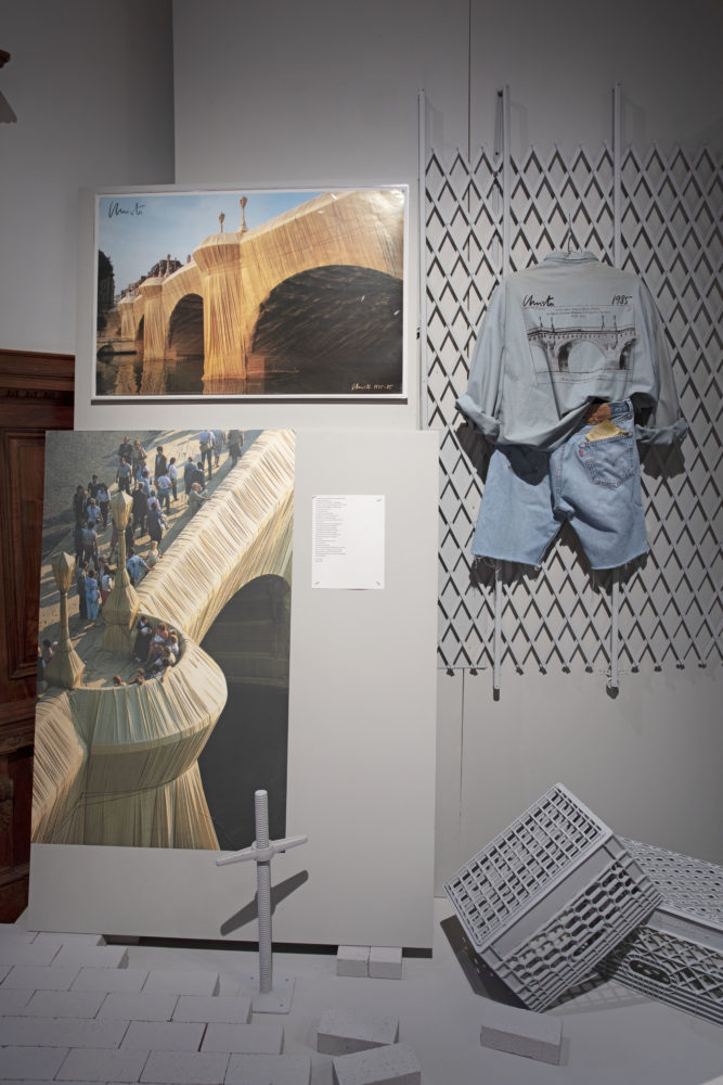 A light blue shirt with a screen-printed image of a bridge displayed on the back. The shirt is paired with jean shorts and hangs from a mounted gray security gate. Beside ensemble are two large printed images displaying a bridge wrapped in gold fabric. A sheet of paper with a printed poem is positioned next to the images. Gray milk crates and bricks are positioned below.