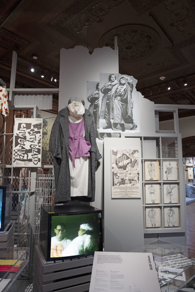 An ensemble hangs against a gray wall with two large overlapping grayscale photographs of models in thick fall clothing to the right. The ensemble is comprised of a white loose-fitting cowl neck dress, paired with a light purple jumper tied at the waist and a charcoal and gray wool coat. To the lower right of the ensemble is a black-and-white poster that reads [SUB-Urban]. Six menswear illustrations are displayed to the right, our right, of the poster.