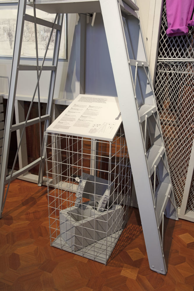 Image displaying a wooden plaque that provides information about the installation and surrounding objects. The plaque is mounted on a gray rectangular cage with a cinder block enclosed. A gray ladder stands above the plaque.