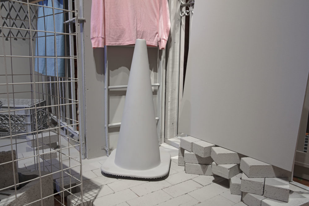 Installation view of a gray traffic cone positioned beside stacked loose gray bricks on a faux paved brick walkway. Part of a gray cage, ladder, and pink long sleeve shirt are captured in the image.