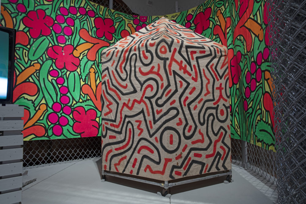 Close-up view of Keith Haring tent for *Secret Pastures* showing black and red line drawing figures seemingly in motion. A brightly colored tarp displaying phallic flowers is behind the tent, hanging across a gray chain-link fence.