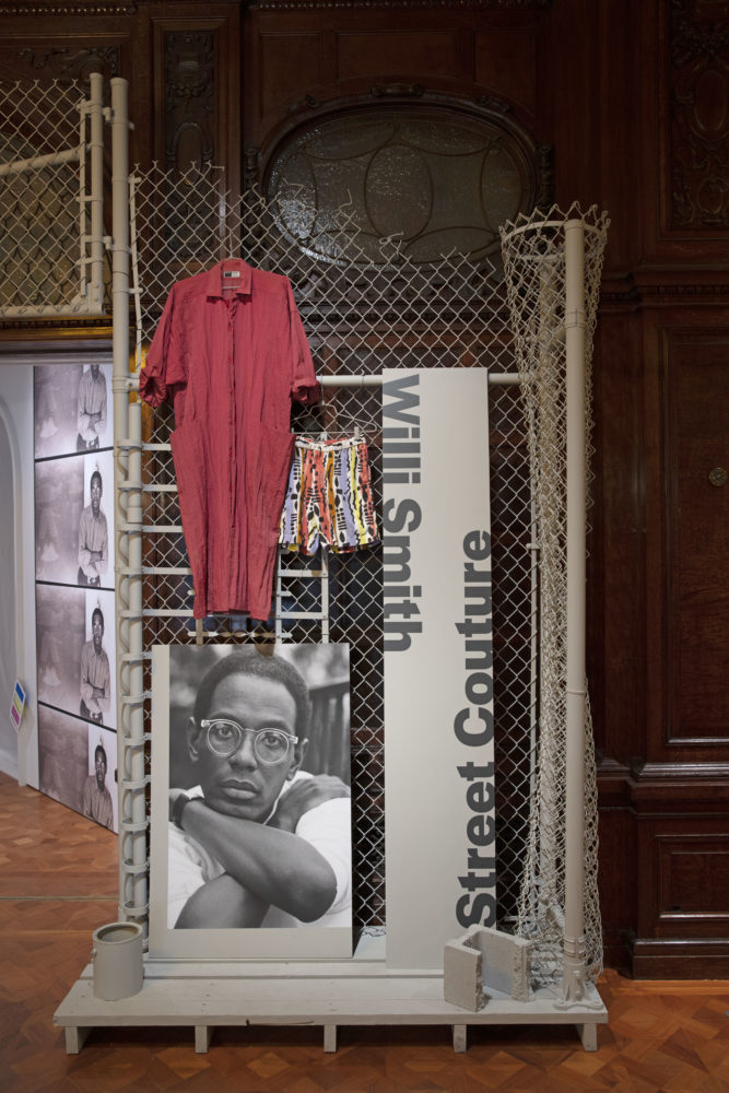 On our right of the entrance in Cooper Hewitt’s Great Hall, a grayscale portrait of a dark-skinned man with round glasses (Willi Smith) rests against a gray metal ladder and chain-link fence. To the right of the portrait, a long board of printed text reads, [/Willi Smith/ Street Couture/]. Beside the board, a pair of multicolored shorts and a long red nylon coat hang from the chain-link fence.