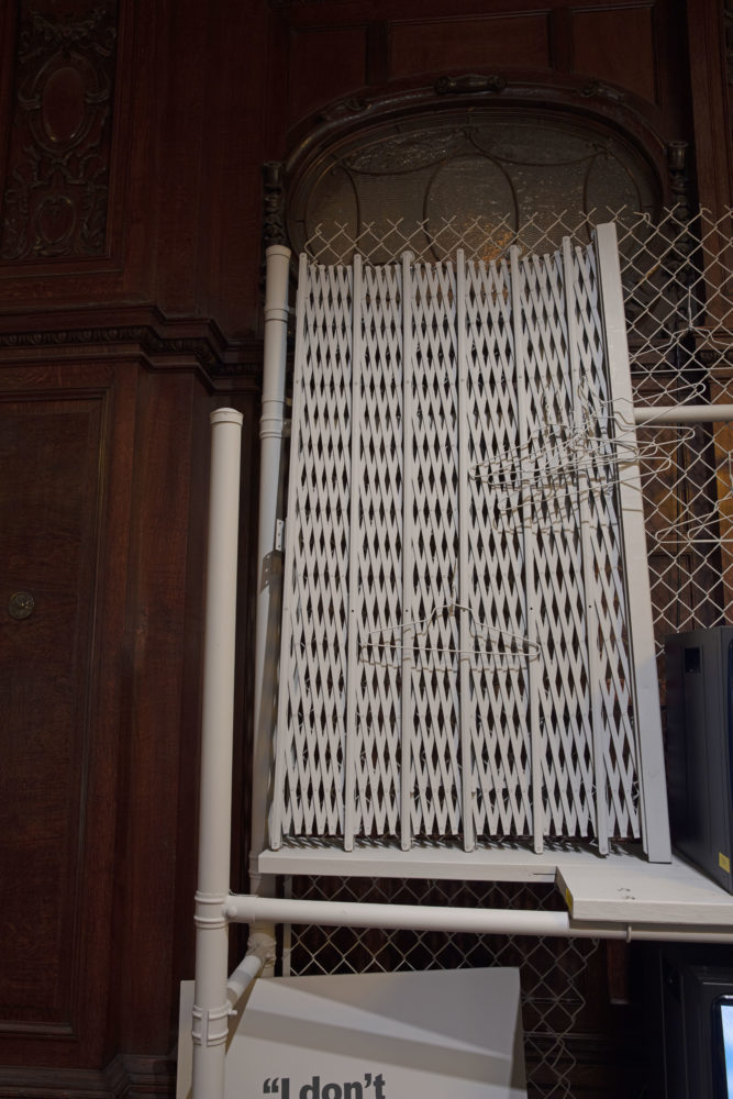 Close-up of a gray folding security gate that sits atop gray wooden planks in a dark wood-paneled room. Gray wire hangers hang from the folded security gate.