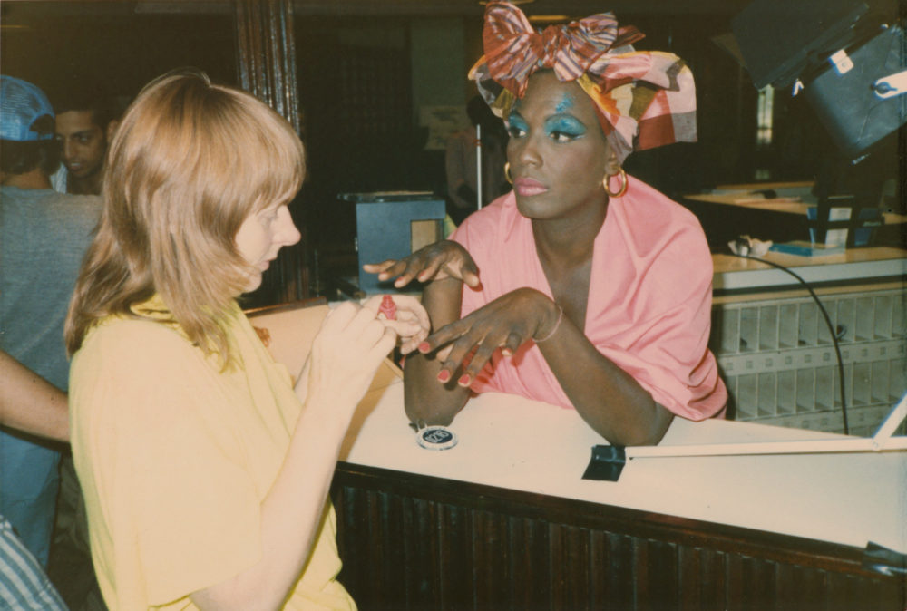 Makeup artist Linda Mason and Willi Smith on the set of Expedition. Willi is dressed in a pink top, and brightly colored bow on his head. He is getting his nails painted and is wearing bright blue eye liner.